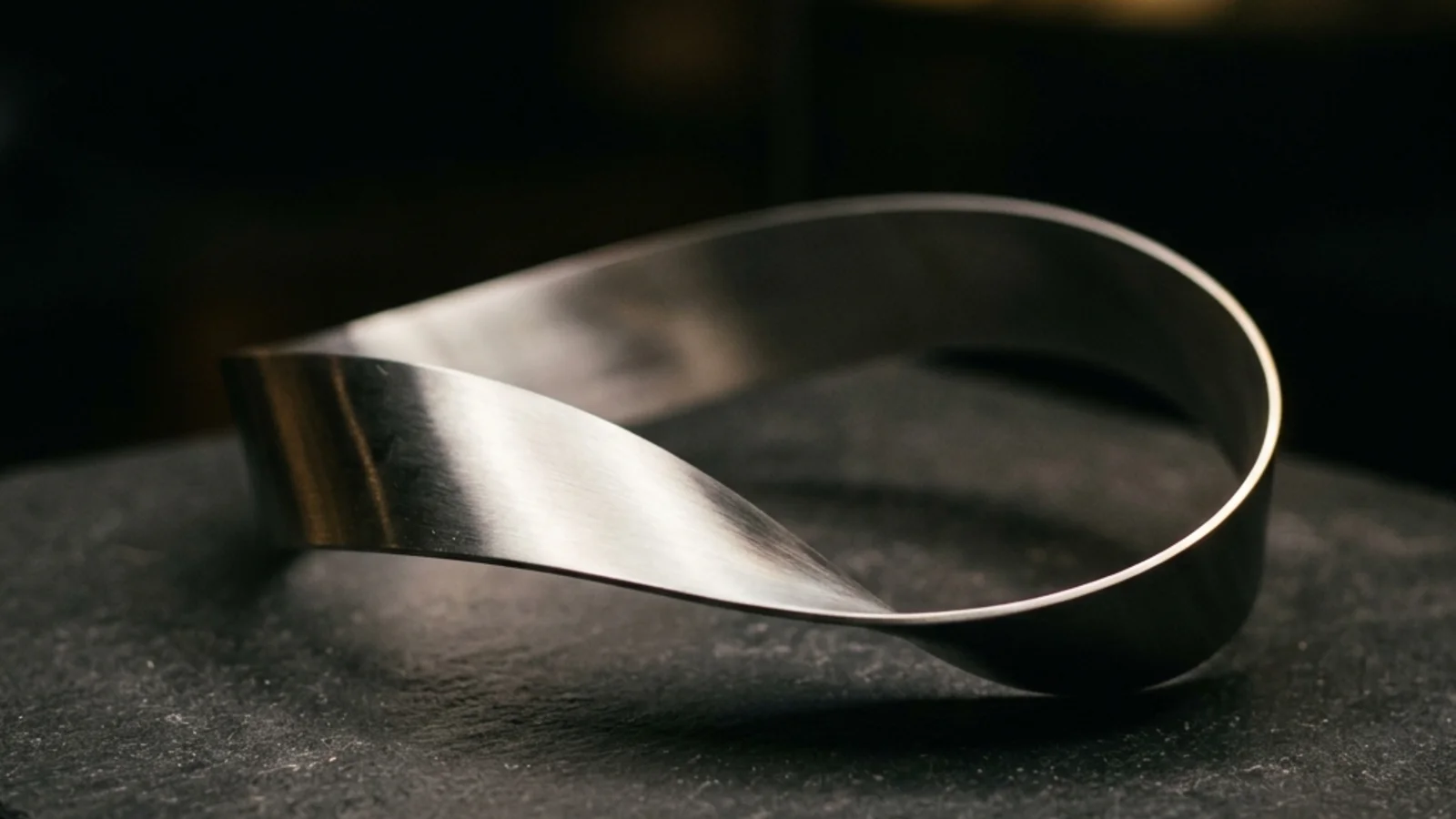 A polished metal Möbius strip resting on a dark surface, with soft directional lighting picking out its single continuous edge that you can trace forever without crossing a boundary